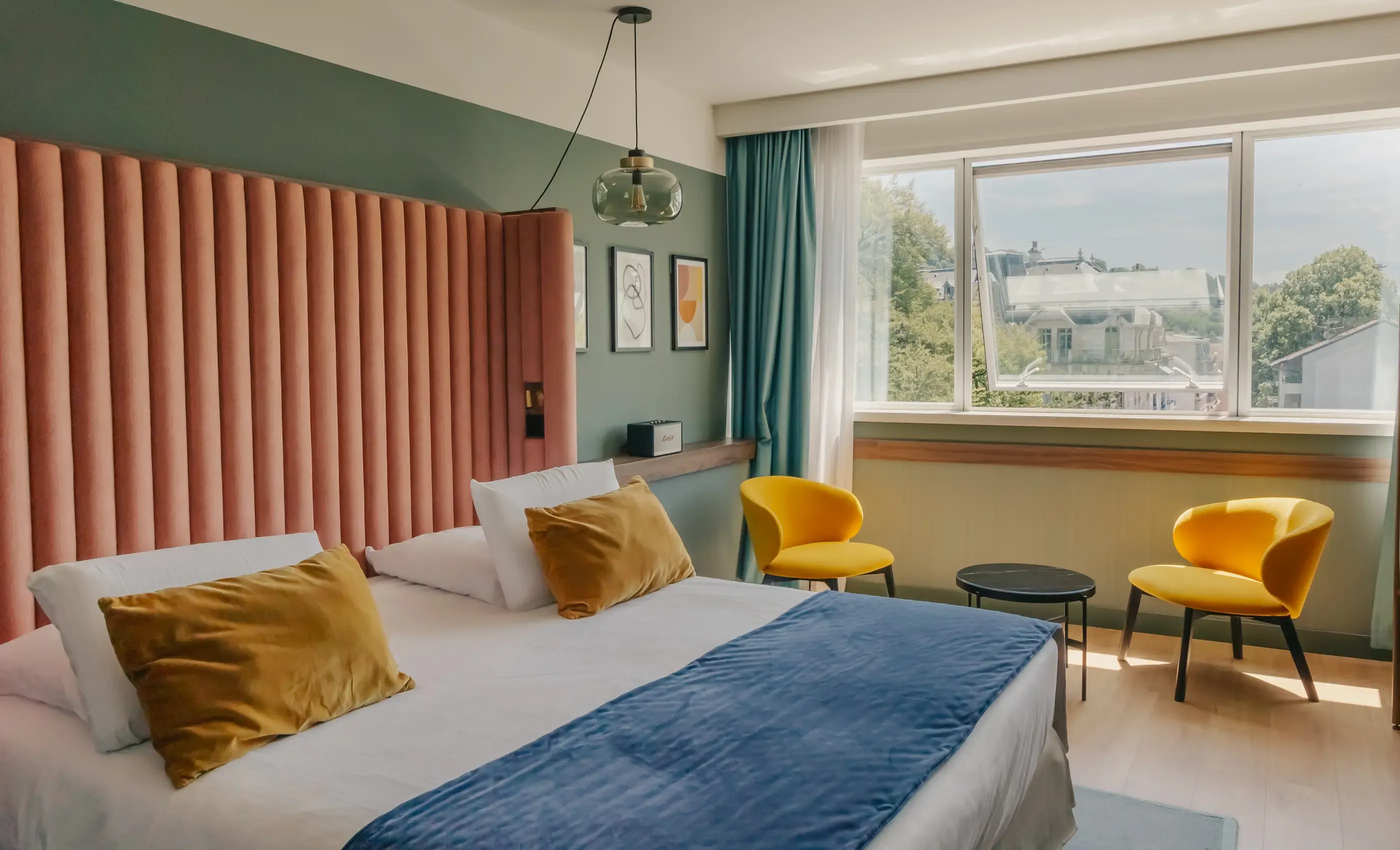 Chambre avec un lit double aux couvertures bleues et blanches, des coussins dorés, deux chaises jaunes, une table basse et une grande fenêtre donnant sur un paysage urbain